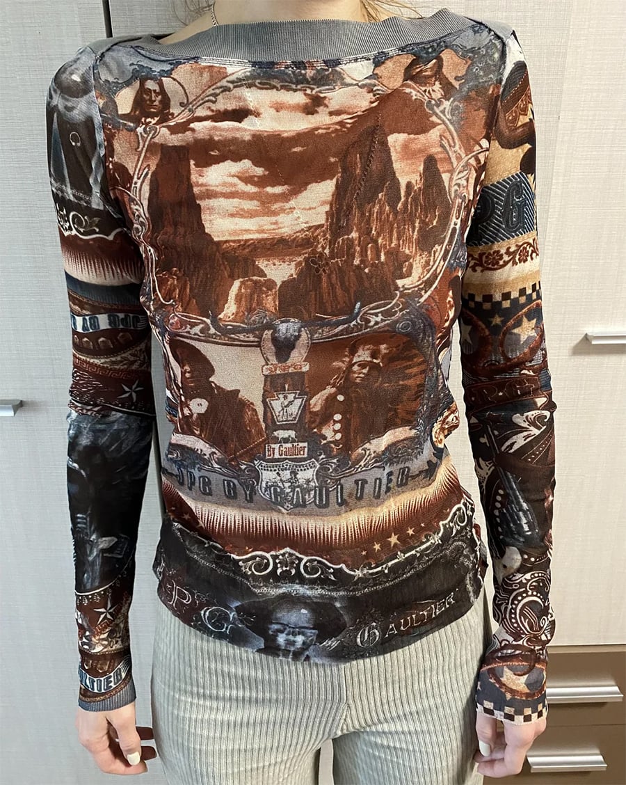 A person wearing a fitted long-sleeve top with intricate, sepia-toned fantasy artwork, including landscapes, faces, and decorative patterns. The photo is taken indoors from the neck to hips.