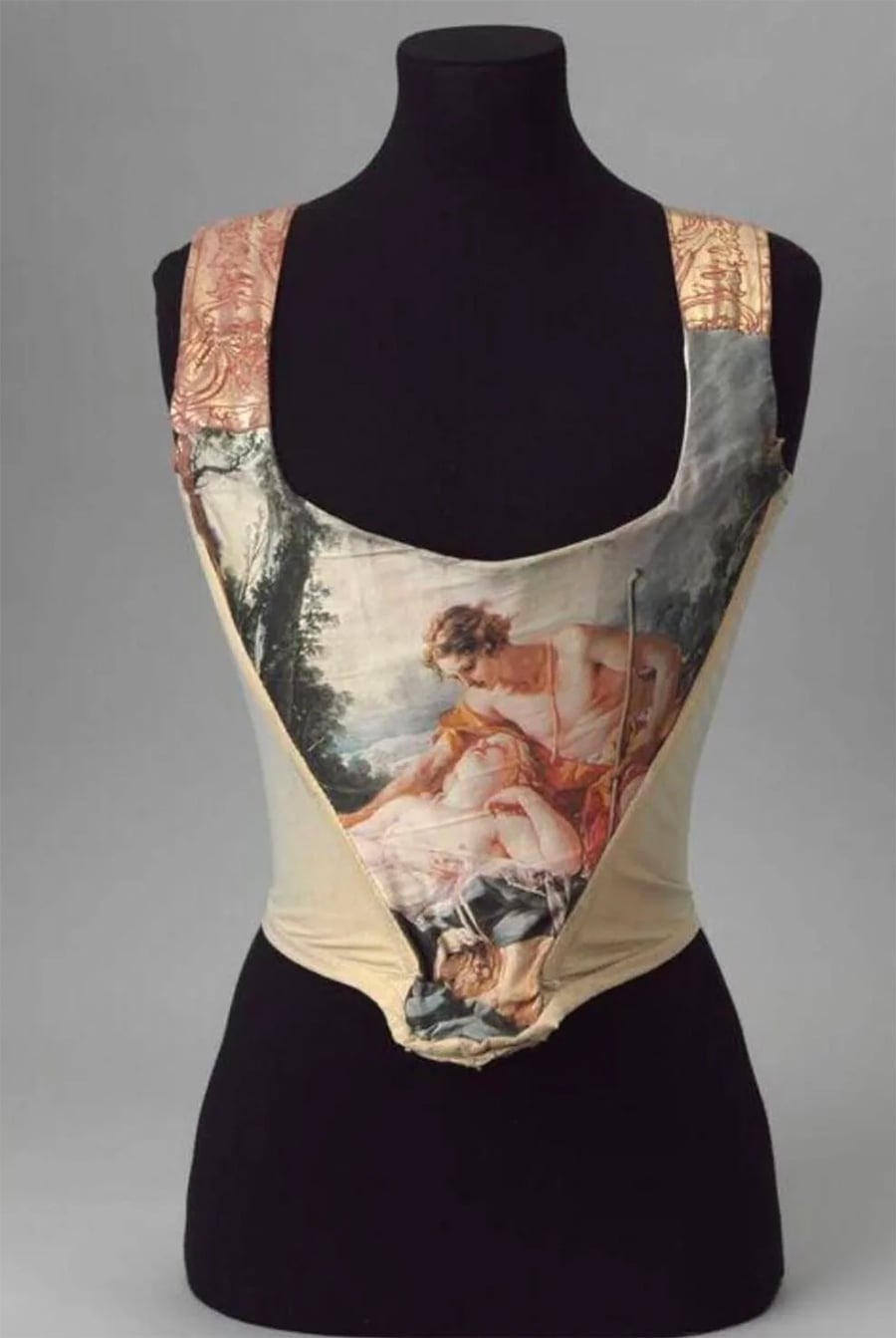 A corset displayed on a black mannequin, featuring a painted scene of two figures in classical attire, bordered by ornate gold and peach fabric on the straps.