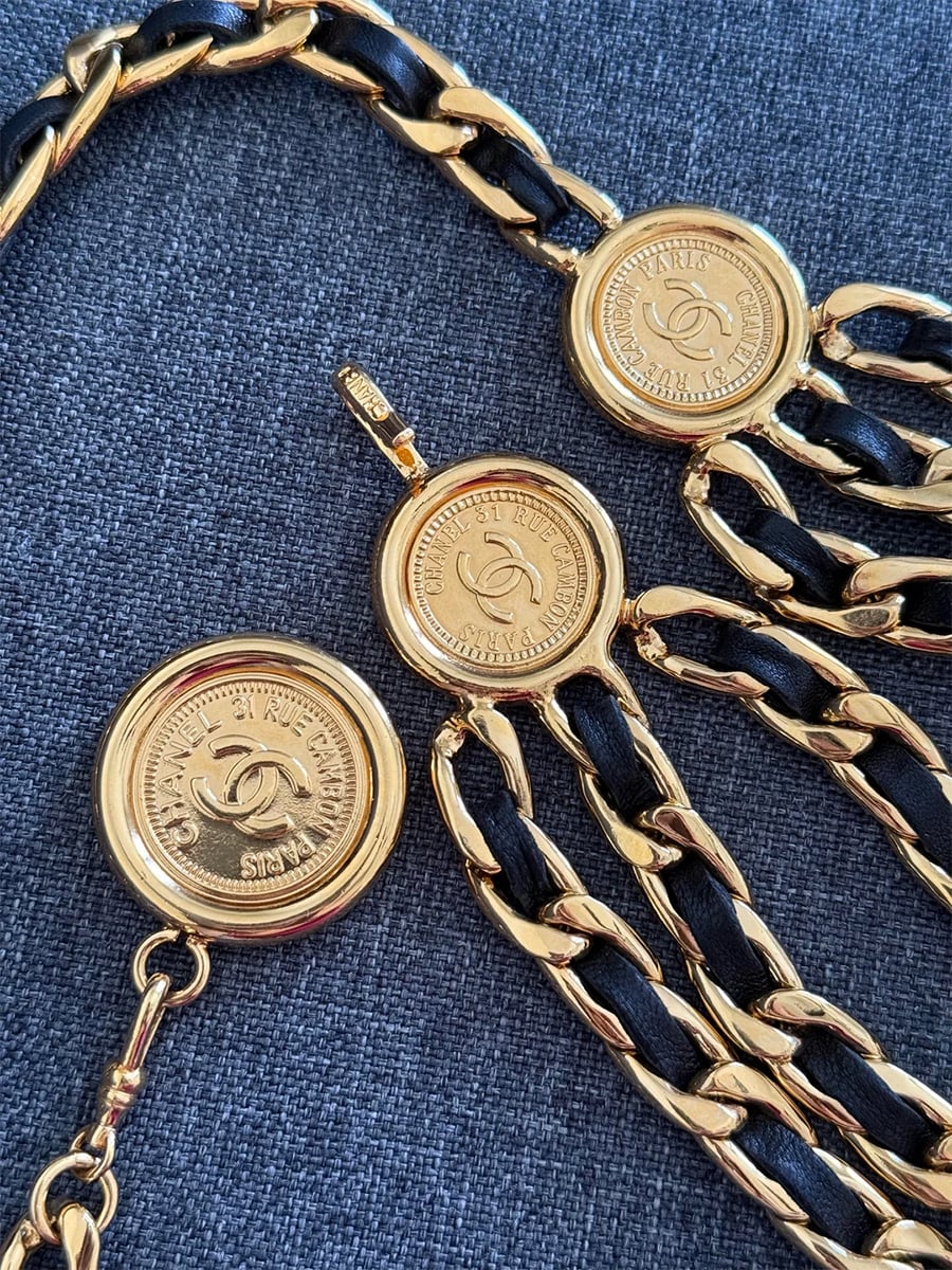 Three gold Chanel medallions with logos, attached to gold and black leather chain links, arranged on a textured gray surface.