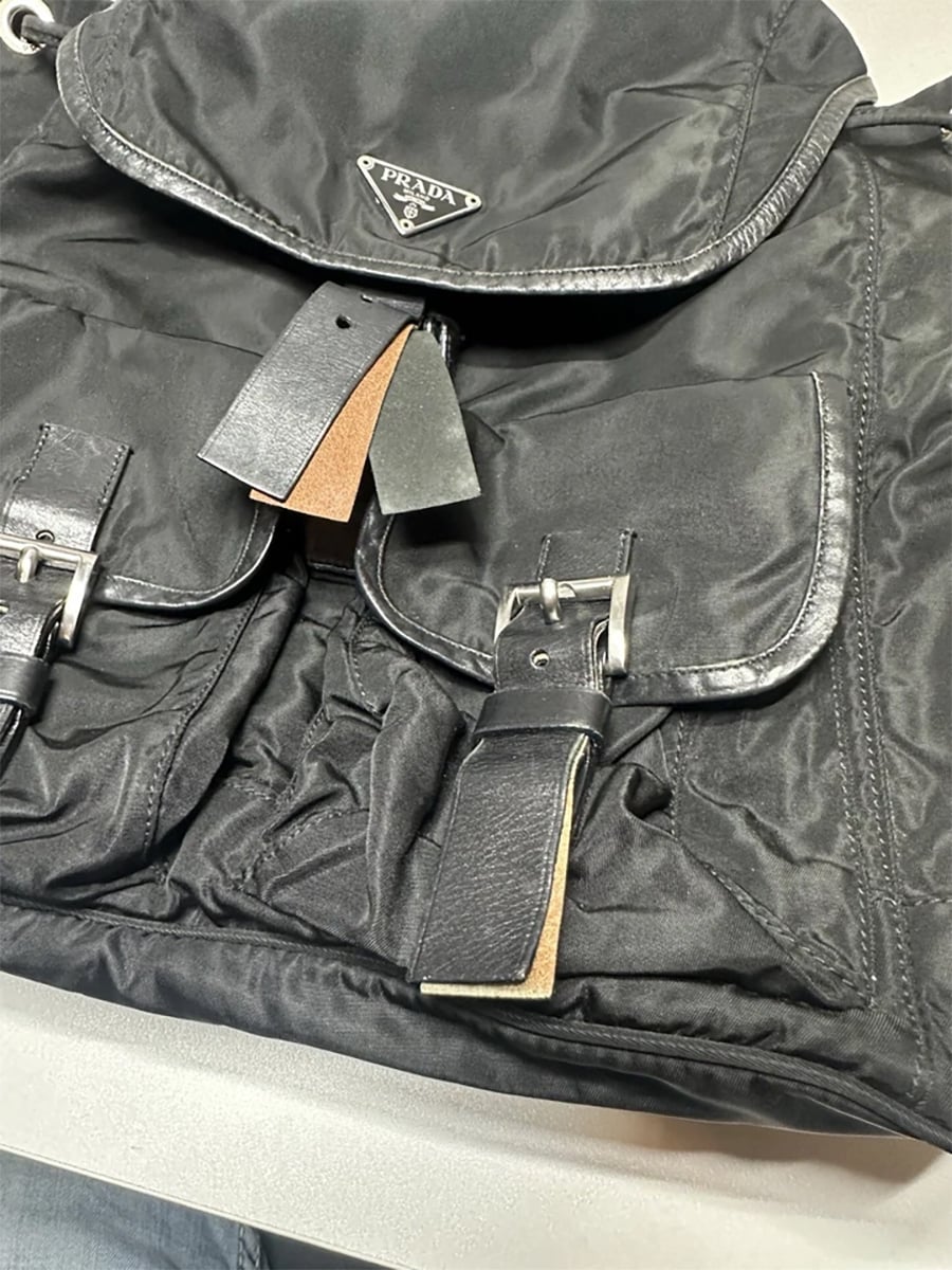 A close-up of a black Prada backpack with two front pockets, silver buckles, leather straps, and the Prada logo visible on the flap. The bag is placed on a light-colored surface.