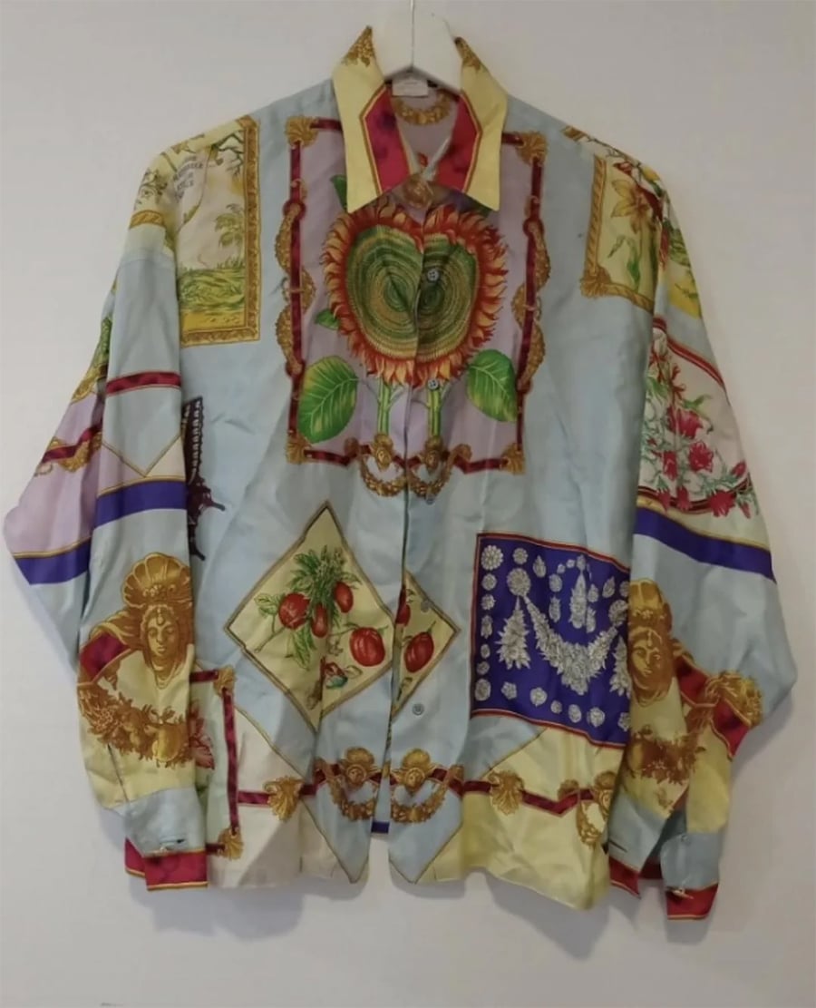 A colorful, long-sleeve button-up shirt with a collage of ornate designs, including sun faces, flowers, hearts, tomatoes, and framed artwork, hanging on a white hanger against a plain wall.