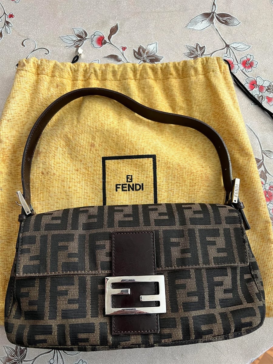 A brown Fendi baguette bag with a black double-F pattern and silver buckle, placed on a yellow Fendi dust bag atop a floral-patterned surface.
