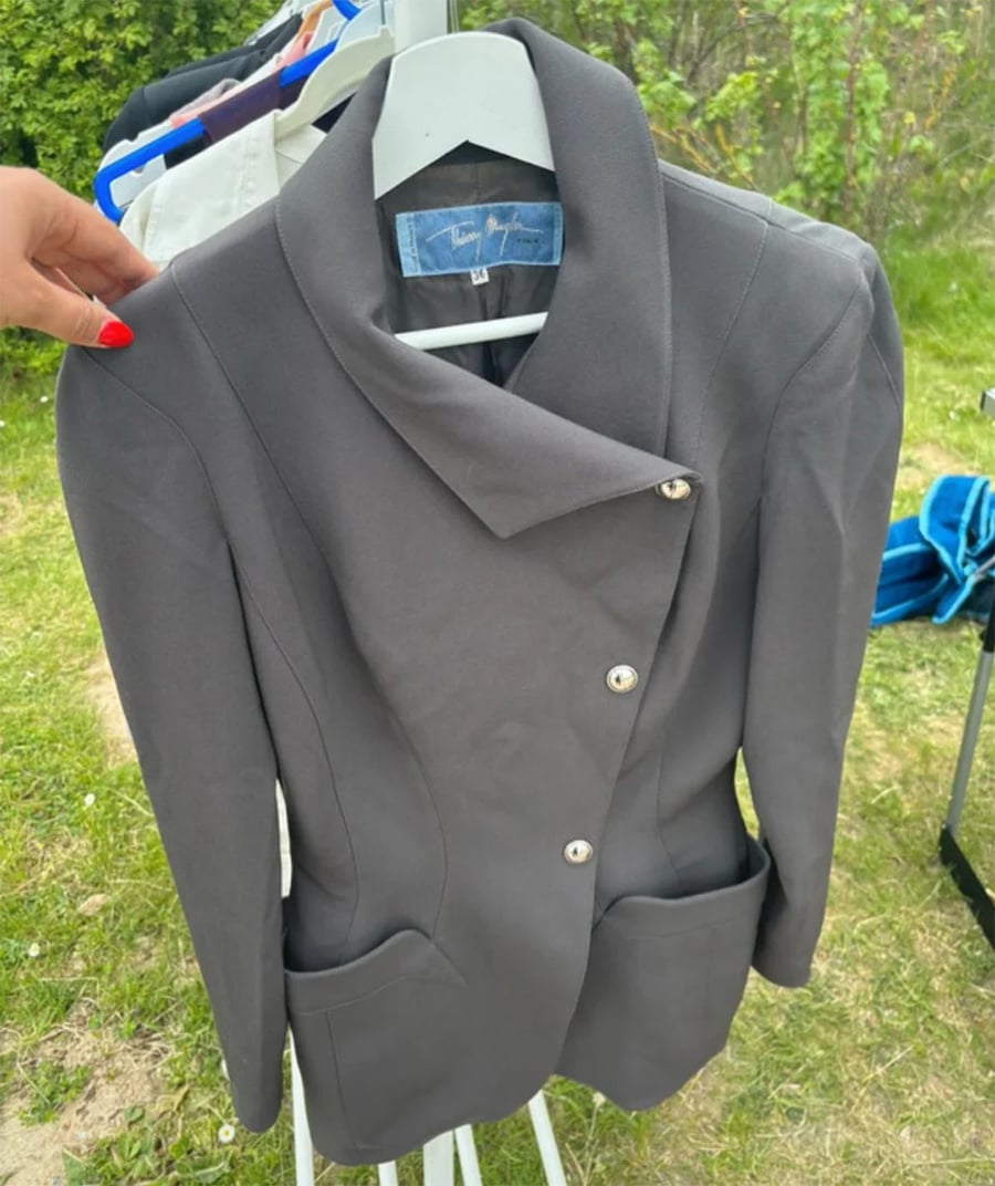 A person holds a gray blazer with asymmetrical buttons, displayed on a white hanger outdoors near greenery.