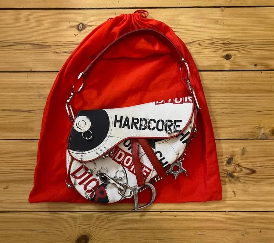 A white Dior saddle bag with bold black and red text, metal charms, and a red strap sits on a red drawstring dust bag atop a wooden surface.