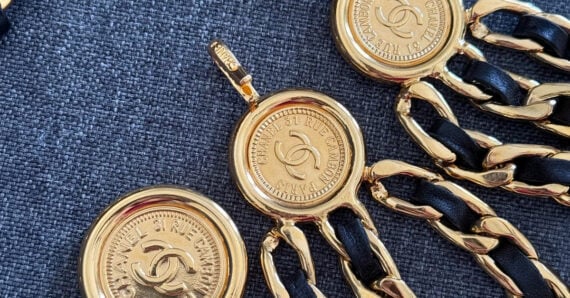 Close-up of gold Chanel jewelry with black leather woven through the chain links, featuring circular pendants stamped with the Chanel logo and branding. The items rest on a textured gray fabric background.