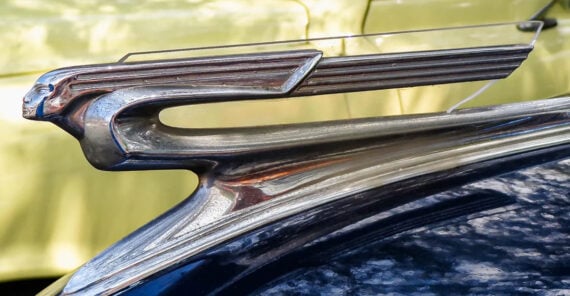 Close-up of a vintage car hood ornament shaped like a stylized, streamlined human figure with wings, set against a blurred yellow background.