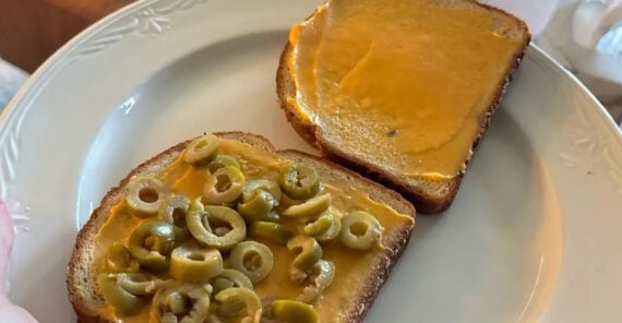 Two slices of bread on a plate, one spread with a yellowish spread and topped with sliced green olives, the other with just the spread.