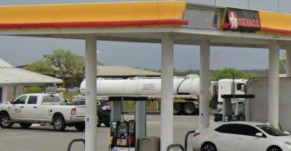 A gas station with a yellow and orange canopy, multiple pumps, a white truck, a white tanker truck, and a silver car parked nearby. Trees and buildings are visible in the background.