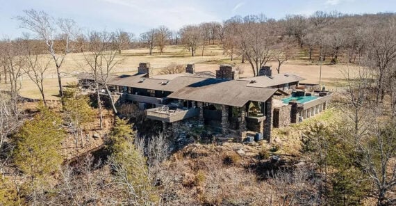 Large modern house with multiple stone chimneys, surrounded by leafless trees and grass fields, viewed from above in a rural setting under a clear sky.