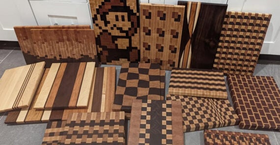 A variety of wooden cutting boards with intricate patterns, including pixel art, stripes, waves, and checkerboards, are displayed against a white kitchen backdrop.
