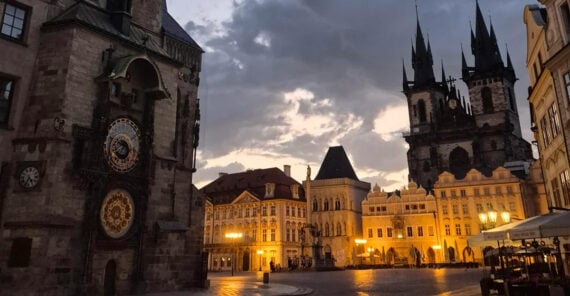 Old Town Square in Prague at dusk, featuring the Astronomical Clock on the left and the Church of Our Lady before Týn in the background, with dramatic clouds and illuminated buildings.