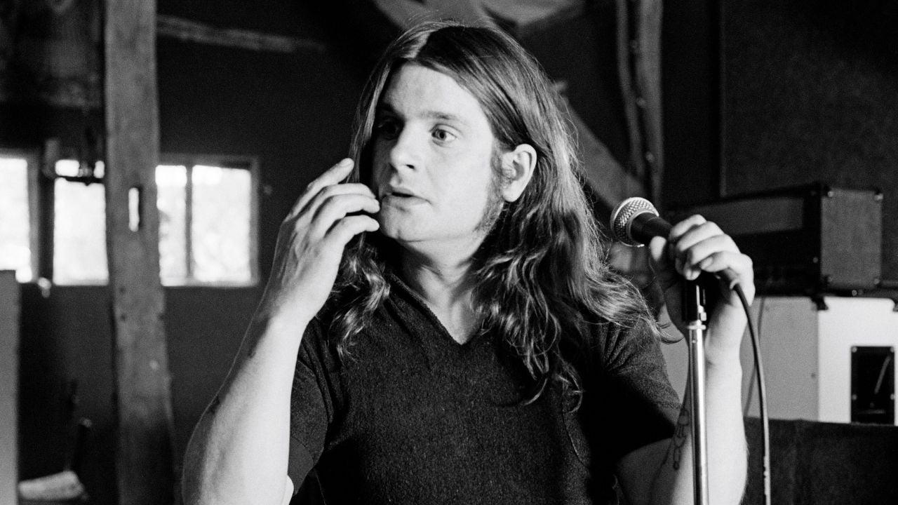 A person with long hair and sideburns holds a microphone and gestures with one hand while looking to the side in a black and white indoor setting.
