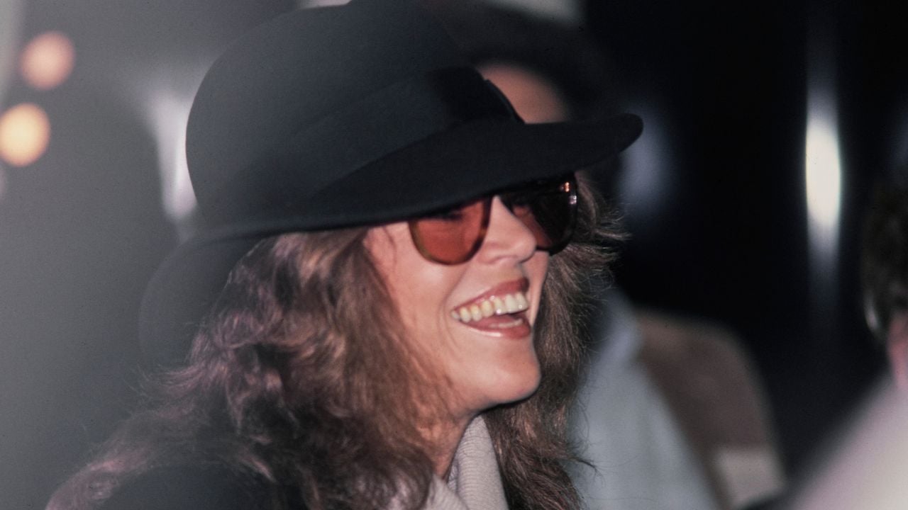 A side-profile shot of a smiling woman with long brown hair, wearing a dark wide-brimmed hat and amber-tinted sunglasses, set against a blurred, dark background.
