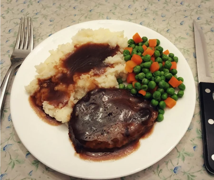 A plate with Salisbury steak in brown gravy, mashed potatoes topped with gravy, and a side of mixed peas and diced carrots. Fork to the left, knife to the right, on a floral tablecloth.