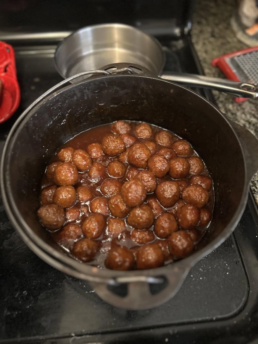 A black pot filled with small meatballs in a thick reddish-brown sauce sits on a stovetop. A metal pot with a ladle is in the background.
