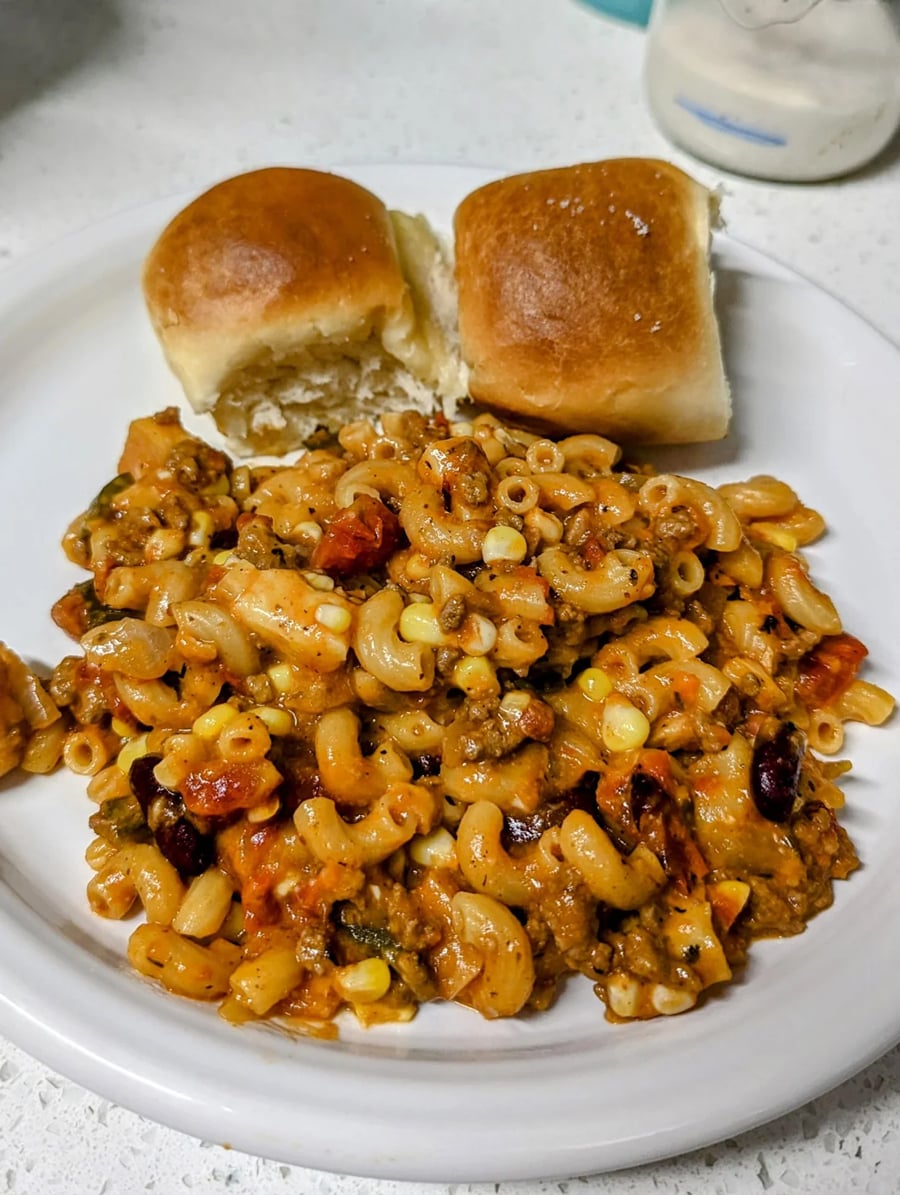 A white plate with macaroni casserole containing ground meat, beans, corn, and tomatoes, served with two soft dinner rolls.