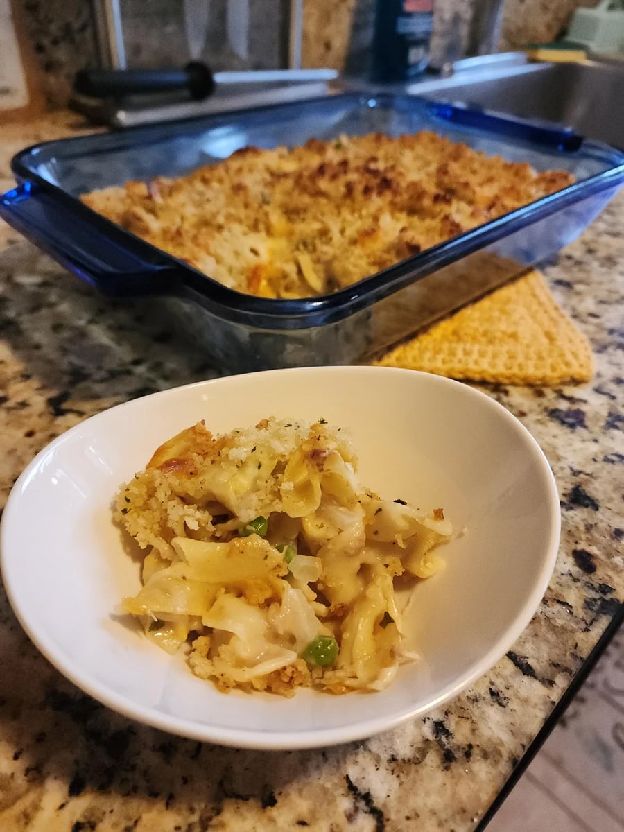 A serving of baked noodle casserole with peas in a white bowl, sitting in front of a blue baking dish of the same casserole on a granite kitchen counter.