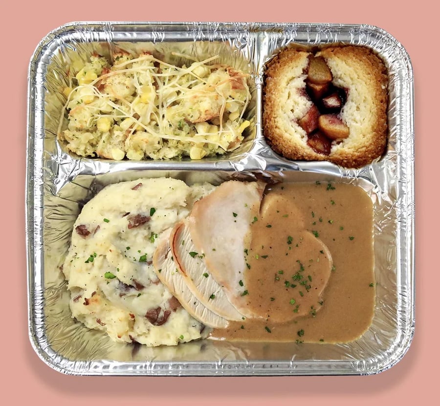 A foil tray with mashed potatoes, stuffing, turkey slices with gravy, and a piece of cake topped with fruit.