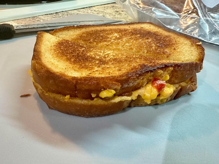 A golden-brown grilled cheese sandwich sits on a cutting board, with melted cheese and small bits of red pepper oozing from the side. A plastic bread bag and knife are in the background.