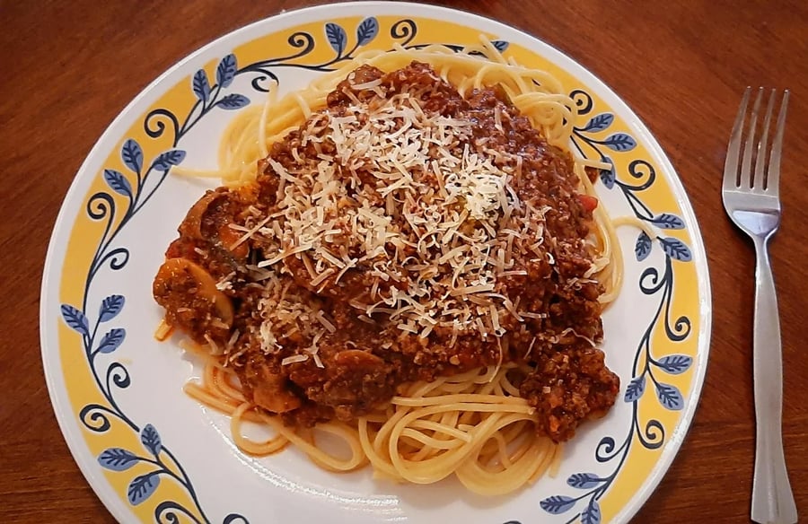 A plate of spaghetti topped with meat sauce and grated cheese is on a decorative plate, with a fork placed to the right on a wooden table.