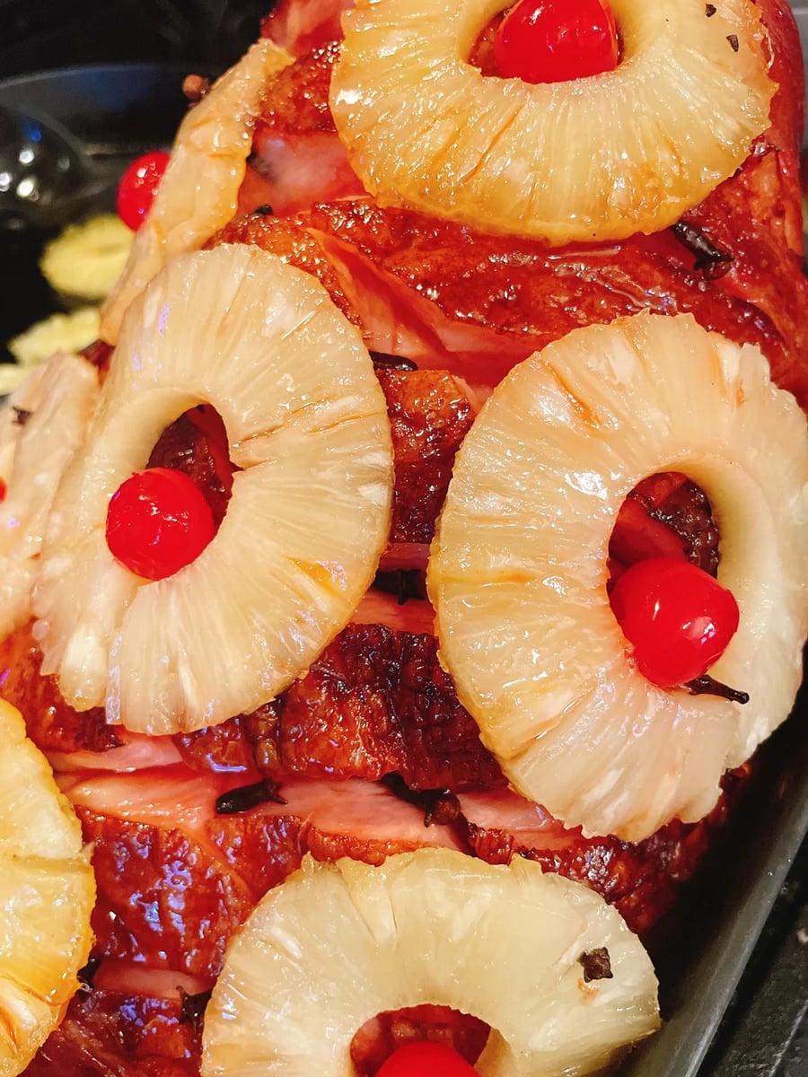Close-up of a baked ham decorated with pineapple rings and maraschino cherries, held in place with cloves. The ham has a glazed, golden-brown exterior and is sliced in a crisscross pattern.