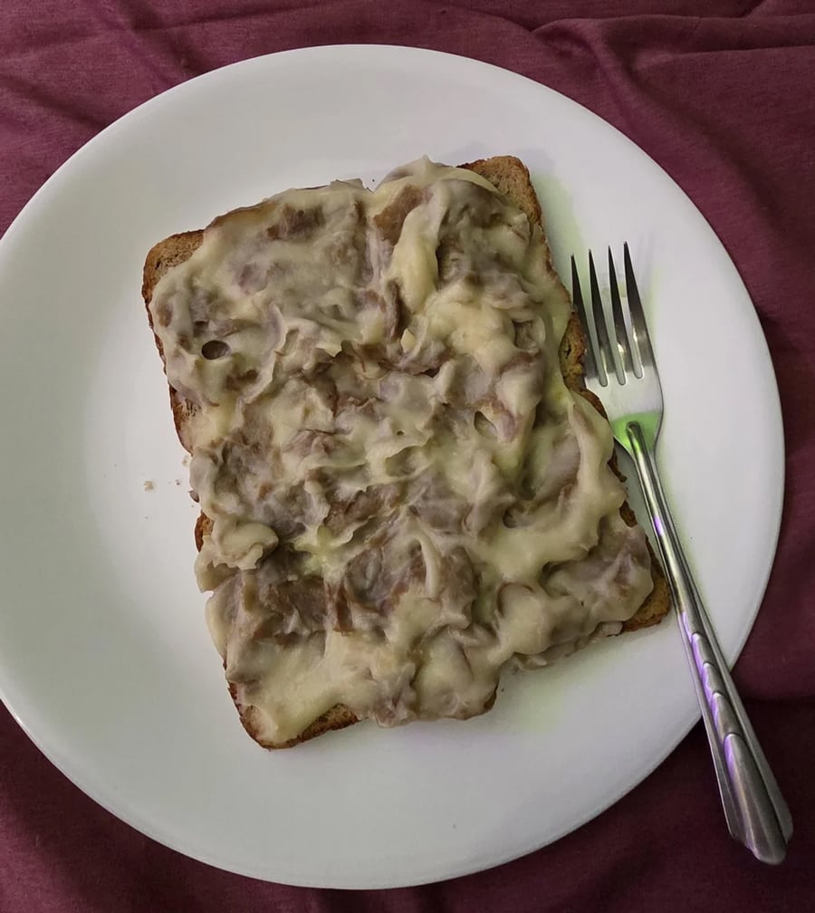 A slice of bread covered with a thick, creamy, grayish topping, served on a white plate with a silver fork on the side. The background is a maroon fabric.