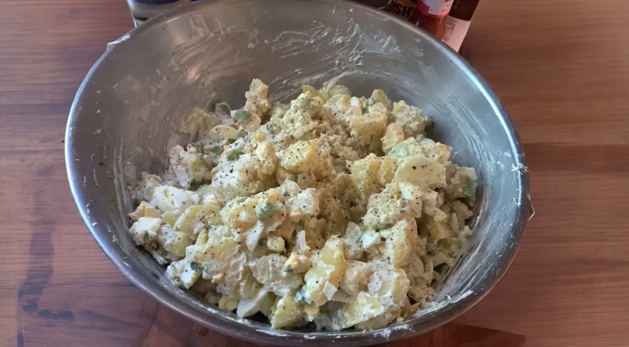 A metal bowl filled with potato salad, containing chopped potatoes, eggs, and herbs, sits on a wooden surface. Seasoning is sprinkled on top. Bottles are visible in the background.