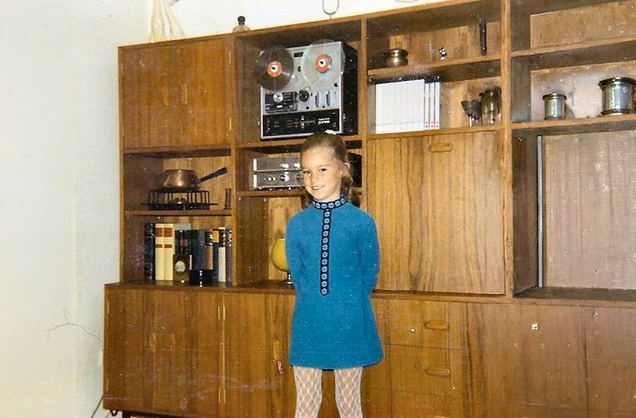 A young child in a blue dress stands in front of a wooden shelving unit with books, pots, and a vintage reel-to-reel tape recorder.