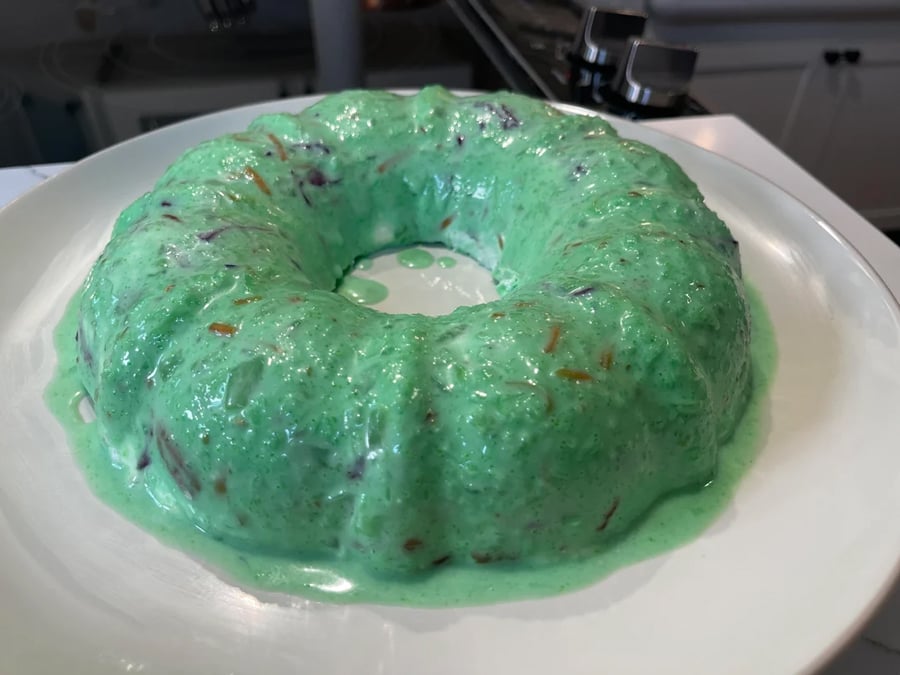 A green gelatin mold with visible bits of fruit, sitting on a white plate in a kitchen setting.