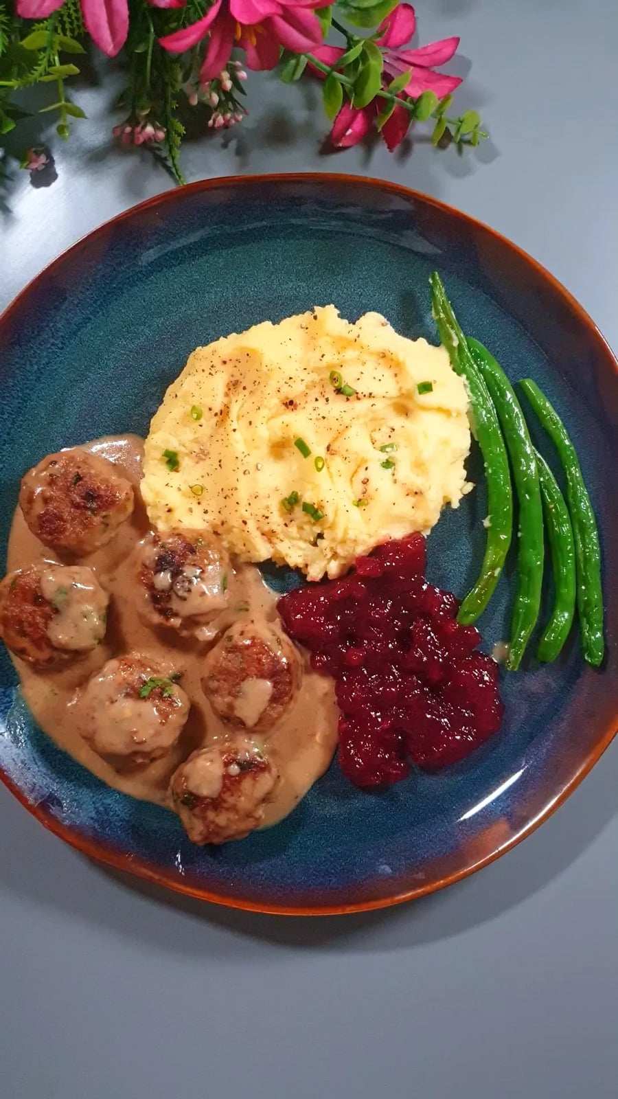 A plate with Swedish meatballs in gravy, mashed potatoes topped with herbs, green beans, and a serving of lingonberry sauce, garnished with chives. Flowers are visible at the top edge of the image.