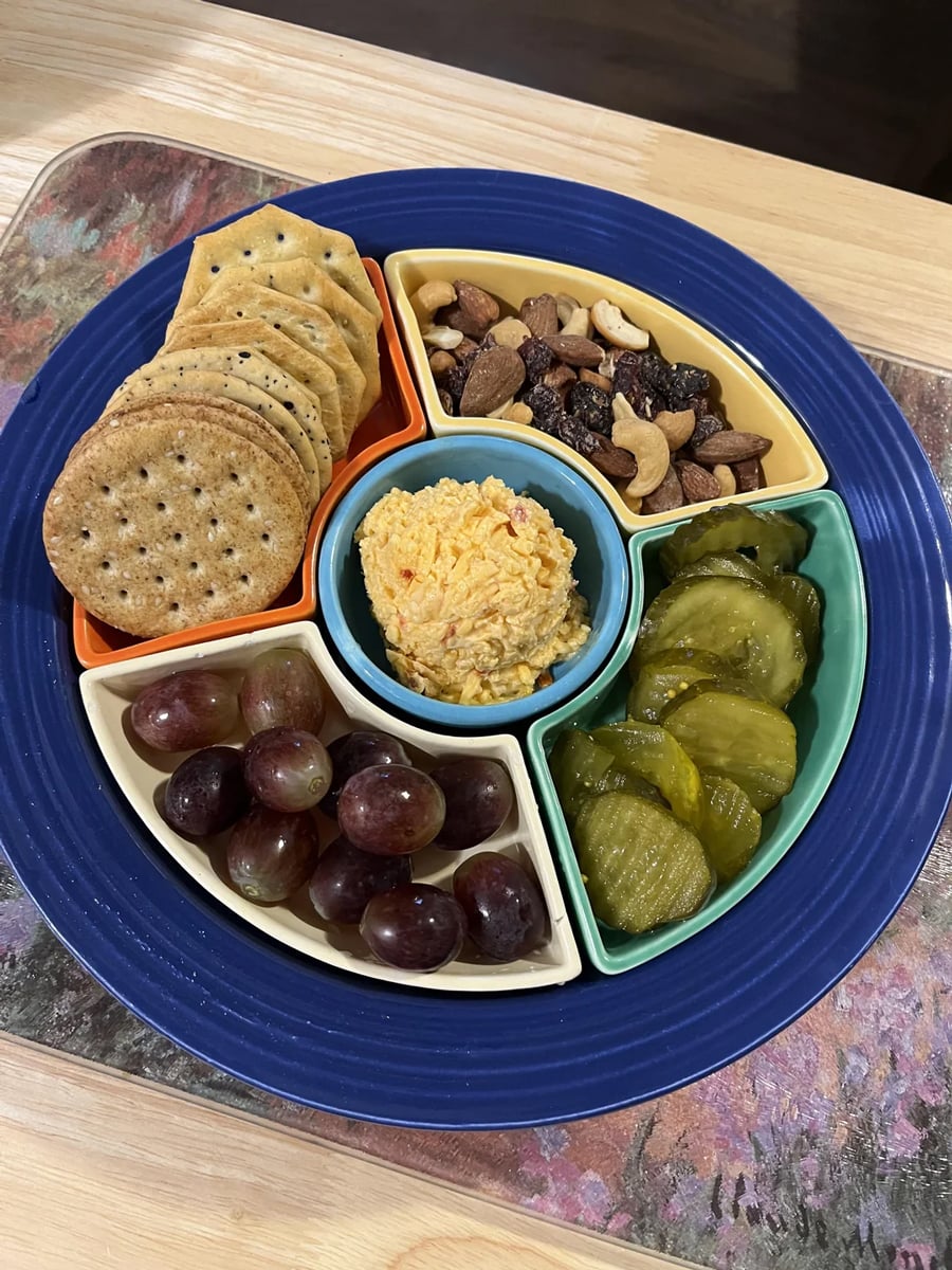 A divided tray with crackers, mixed nuts, pickles, grapes, and pimento cheese spread in the center, arranged on a blue plate on a wooden table.