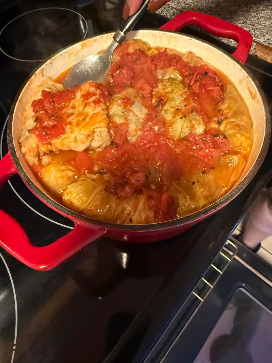 A red pot on a stove filled with cabbage rolls topped with tomato sauce, some partially scooped with a spoon.
