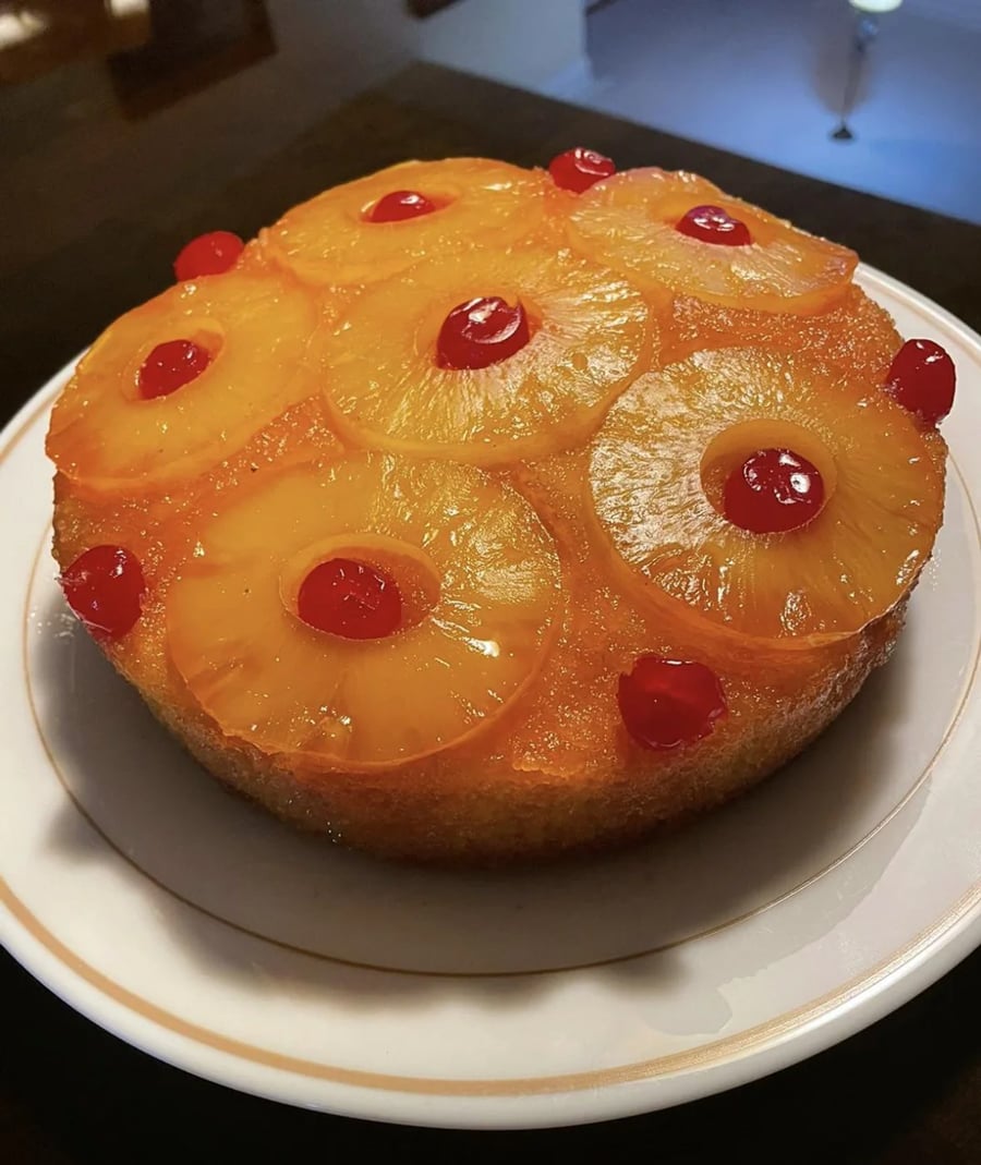 A round pineapple upside-down cake topped with pineapple rings and red cherries is displayed on a white plate with a gold rim.