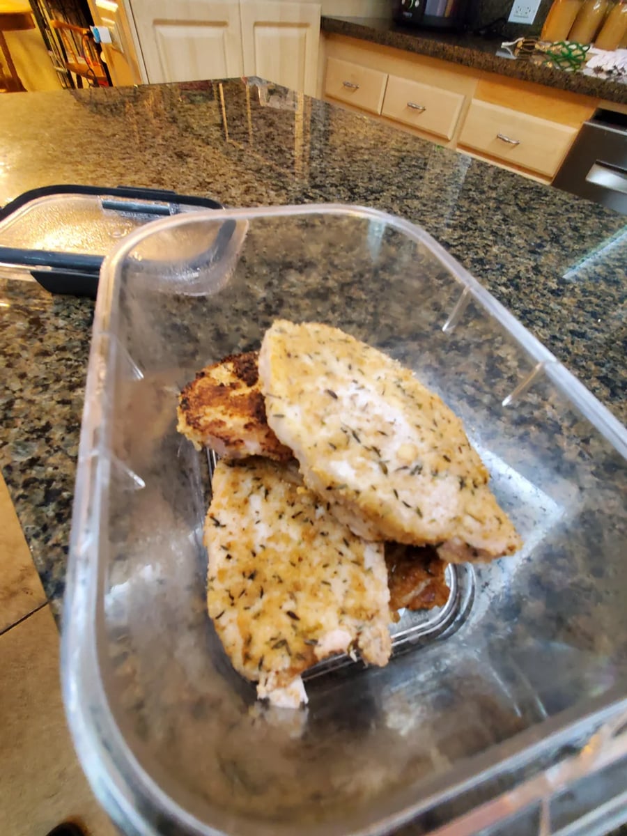 Three cooked, seasoned chicken breasts in a clear plastic container sit on a granite kitchen countertop. The kitchen background includes wooden cabinets and a stainless steel dishwasher.