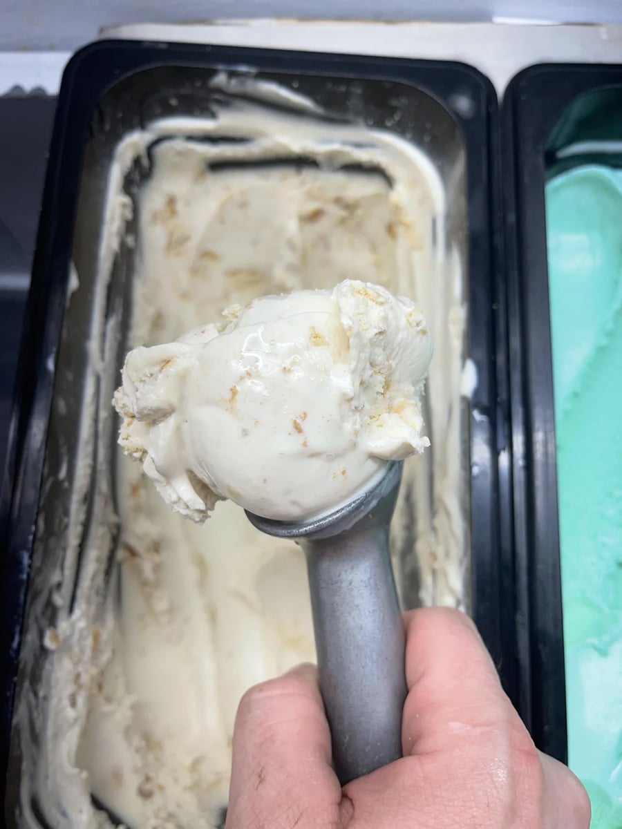 A hand holds an ice cream scoop with a ball of creamy white ice cream above a partially empty container; mint green ice cream is visible in a neighboring container.