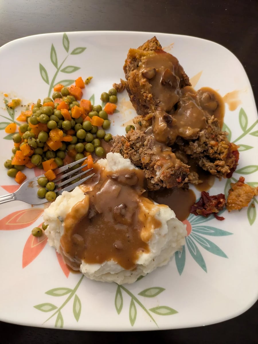 A plate with meatloaf topped with brown gravy, mashed potatoes with gravy, and a side of peas and diced carrots. A fork rests on the plate. The plate has a leaf pattern design.