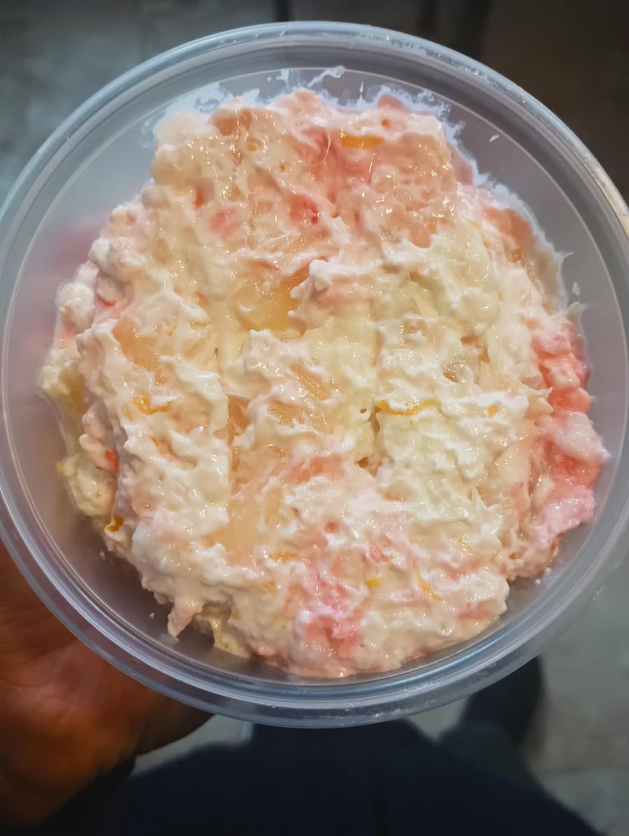 A hand holds a round plastic container filled with creamy fruit salad, featuring a mix of pink, white, and yellow chunks, likely fruit pieces in a thick, milky dressing.