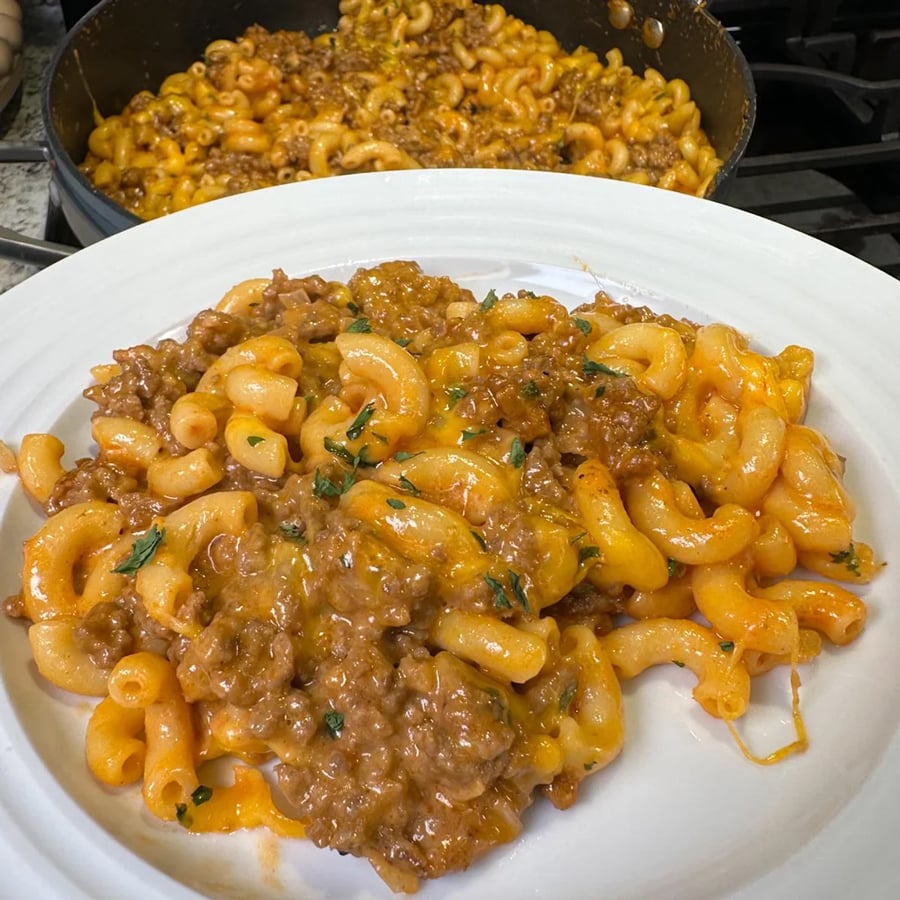 A white plate with creamy, cheesy macaroni and ground beef, garnished with herbs. A pan filled with more of the same pasta dish is in the background on a stove.