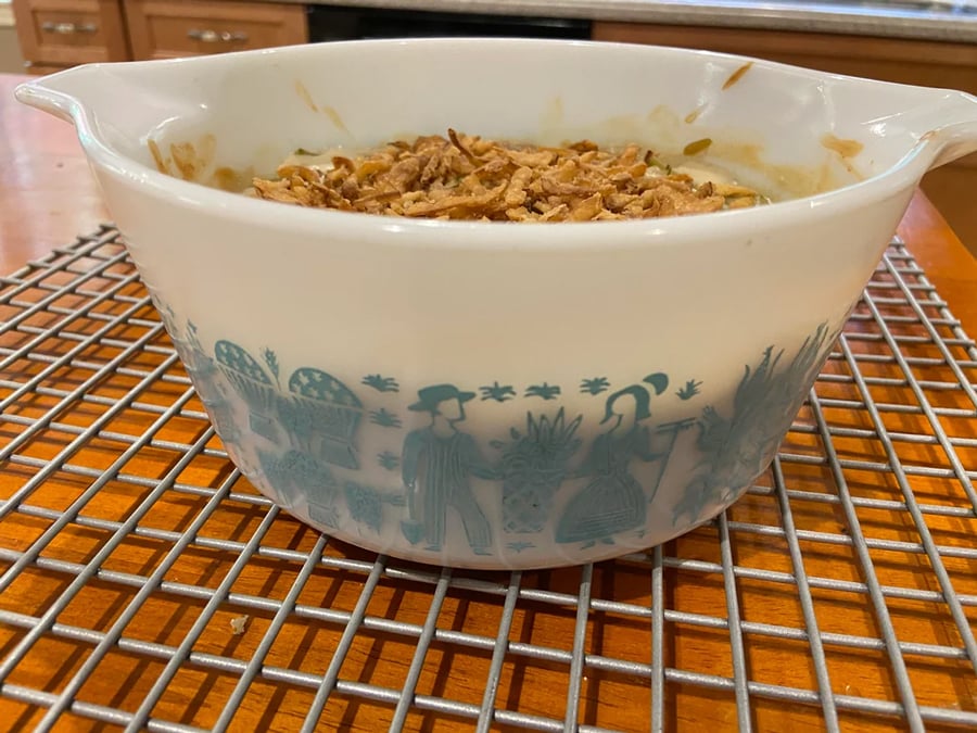 A vintage Pyrex bowl with blue farmer and farm designs, filled with a casserole topped with fried onions, sits on a metal cooling rack on a wooden table.