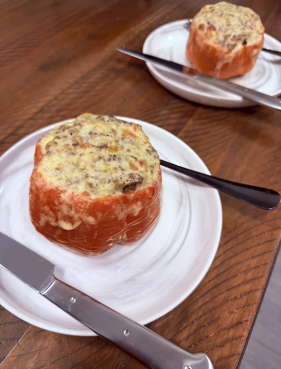Two white plates each hold a stuffed tomato topped with melted cheese. Knives and forks are placed on the plates, and everything rests on a wooden table. The dish appears hearty and savory.