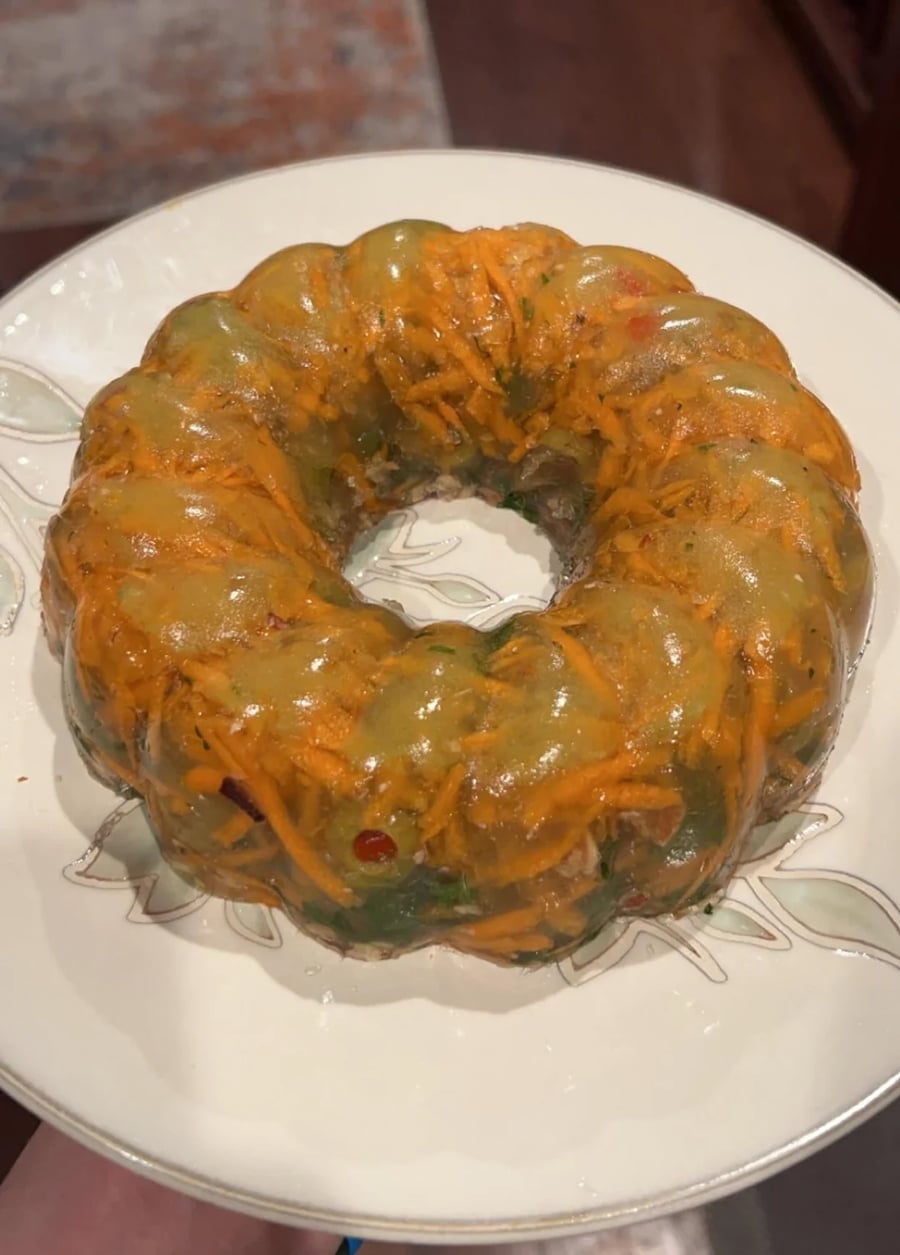 A clear gelatin ring mold with shredded carrots and bits of green and red vegetables, served on a white plate with a leaf design.