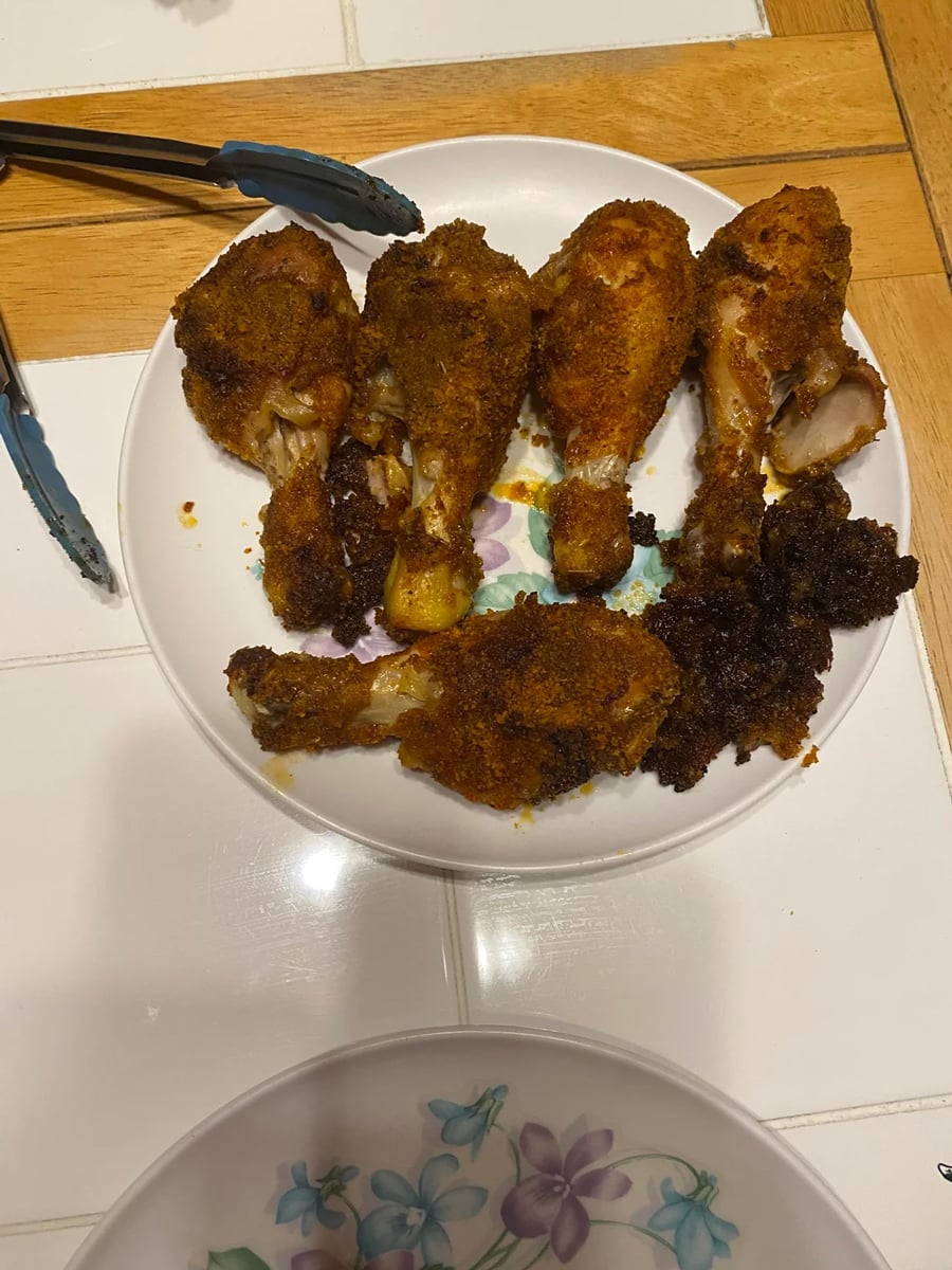 A plate with five breaded and baked chicken drumsticks, arranged on a white plate with a floral design. A pair of tongs and a bowl with a flower pattern are nearby on the wooden and tiled countertop.