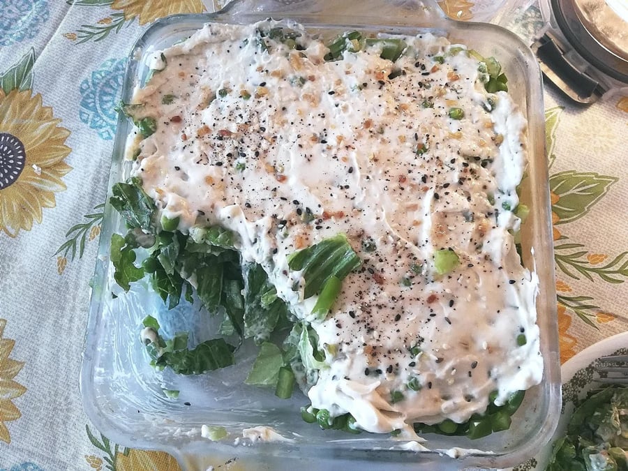 A glass dish with a layered salad topped with creamy dressing and cracked pepper, with a large scoop missing from one corner. The salad sits on a sunflower-patterned tablecloth.