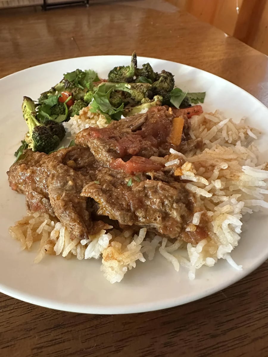 A plate of white rice topped with saucy beef curry, served with a side of roasted broccoli and mixed vegetables on a wooden table.