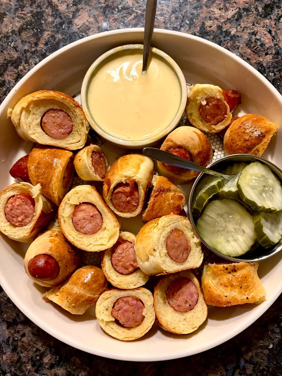 A plate with pigs in a blanket, a bowl of cheese dip with a spoon, and a side of sliced pickles on a granite countertop.