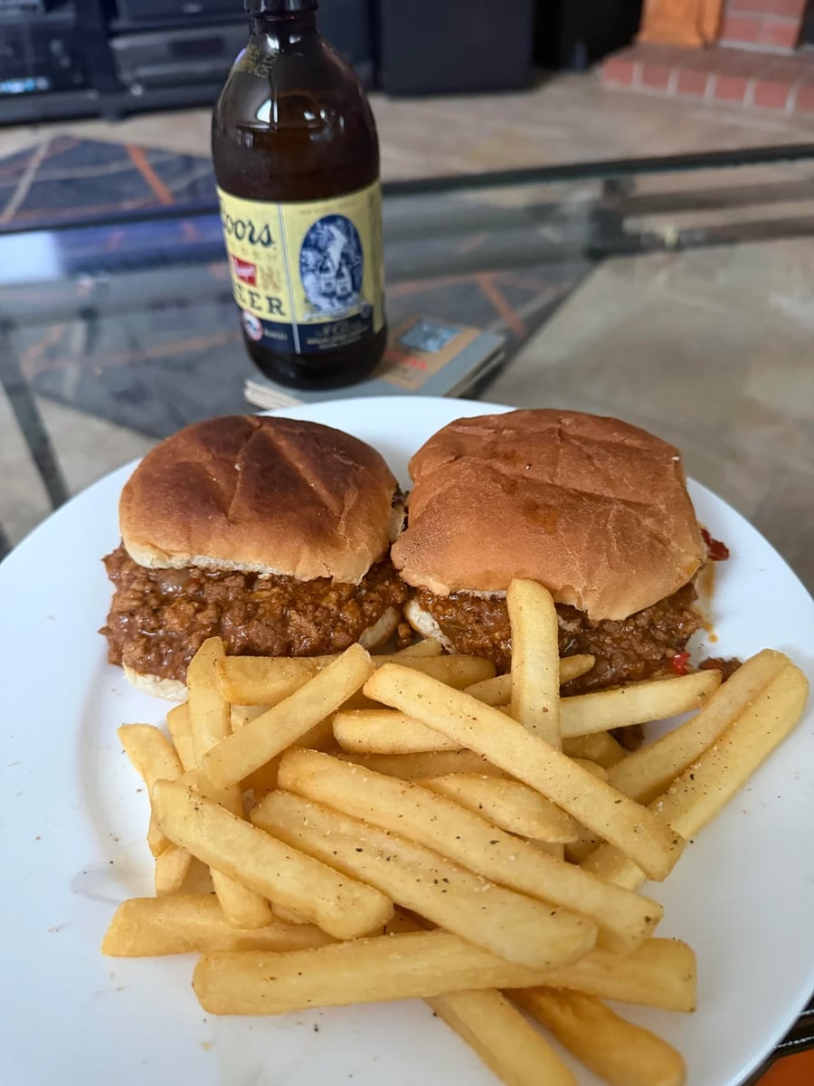 A white plate with two sloppy joe sliders and a serving of seasoned French fries sits on a glass table. Behind the plate is a bottle of beer.