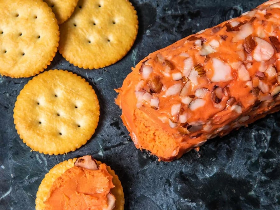 Round crackers and a log of orange cheese spread coated with chopped nuts are on a dark surface. One cracker is topped with the cheese spread.