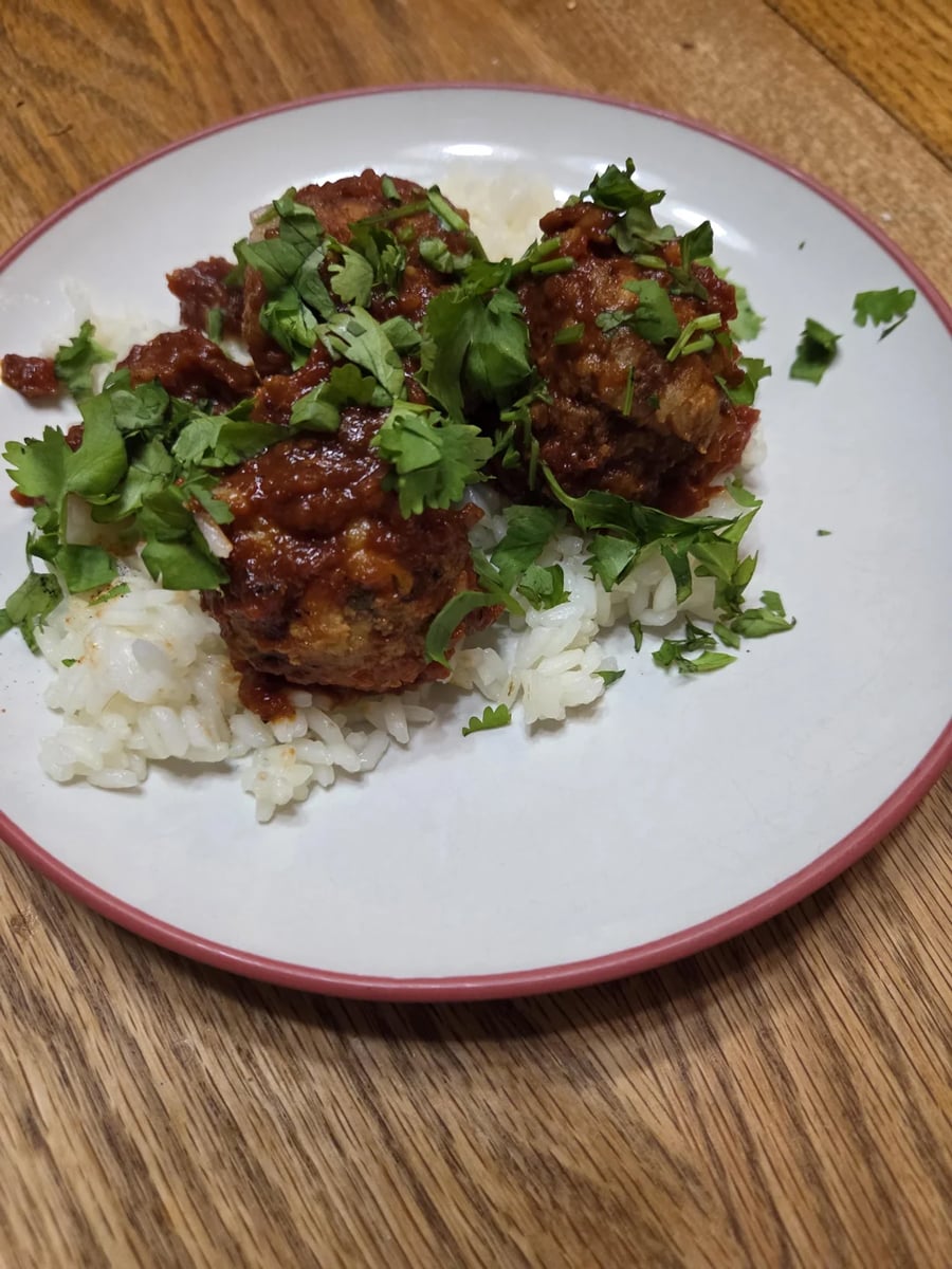 Three meatballs in tomato sauce sit on a bed of white rice, garnished with chopped fresh cilantro, served on a white plate with a pink rim on a wooden table.