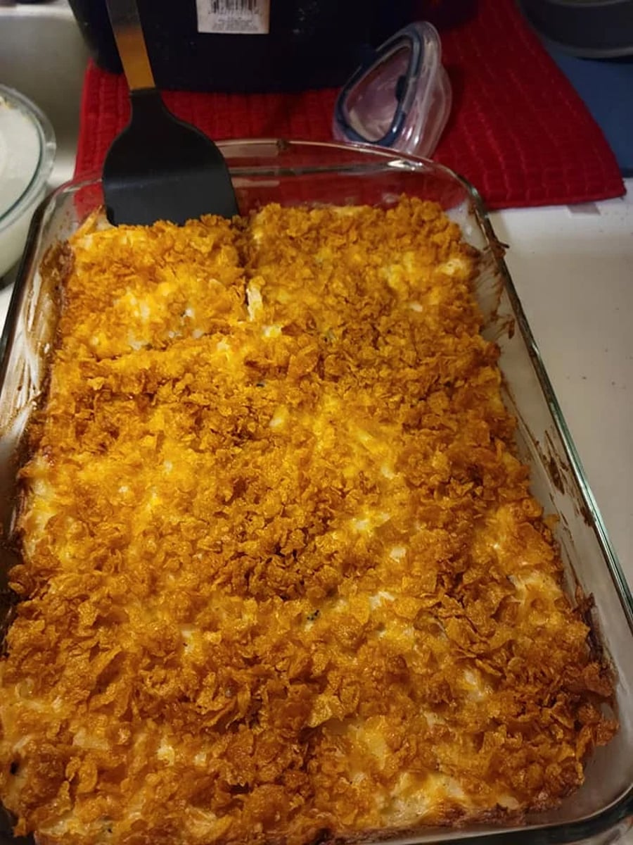 A glass baking dish filled with a baked casserole topped with a layer of crispy, golden-brown cornflakes. A black spatula rests in the dish, and some portions appear to be cut.
