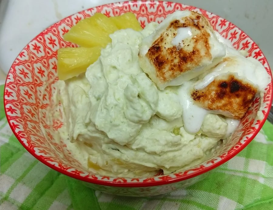 A patterned bowl with creamy green dessert, pineapple chunks, and toasted marshmallows on top, sits on a green-checkered cloth.